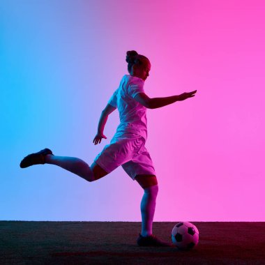 Dişi futbolcu, dinamik bir tekme atıyor, bacağını kaldırıyor ve pembe mavi arka planda neon ışıkları altında keskin bir odak noktası oluşturuyor. Spor, enerji, rekabet, hareket, eğitim kavramı
