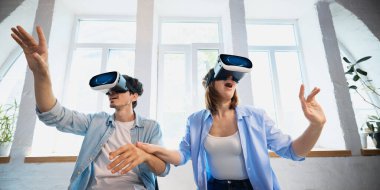 Young couple experiencing virtual reality with VR headsets in a bright, modern living room. Concept of immersive technology enhancing home entertainment experiences.