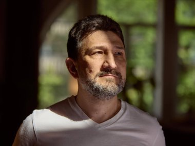 Duygusal derinlik. Beyaz tişörtlü beyaz bir adam, sıcak bir odadaki güneşli pencereden dikkatlice bakıyor. Huzurlu bir ortamda içgözlem ve tefekkür kavramı.