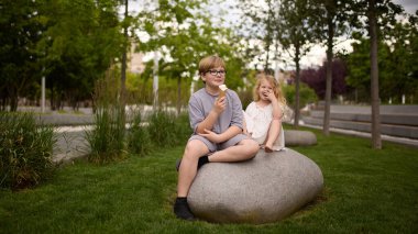 Oğlan ve kız dondurma yiyorlar, yaz parkında taşın üzerinde oturuyorlar. Kardeş bağı, çocukluk mutluluğu, doğa istirahatı, aile gezisi, açık hava tatlısı, masum gülümseme, kaygısız zaman..