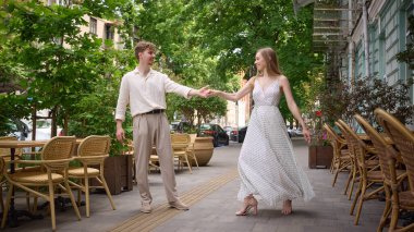 Yeşil ve kentsel geçmişi olan rahat bir kafe terasında dans eden mutlu genç çift. Yaşam tarzı, romantizm, neşe, gündelik zarafet, şehir eğlencesi ve bağlantı kavramı.