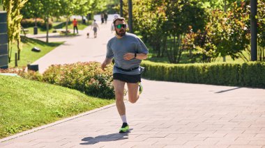Güneş gözlüklü ve kulaklıklı bir adam güneşli parkta koşuyor. Kendine güven temposu, kentsel zindelik, erken çalışma enerjisi, açık hava hareketi ve sağlıklı ritim kavramı.