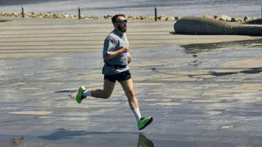 Orta yaşlı koşucu kılık kıyafetiyle güneşli sahil boyunca koşar. İstikrarlı hareket kavramı, kentsel akış, somut ritim, kardiyo odağı ve liman enerjisi..