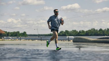 Bulutlu mavi gökyüzünün altında güneş gözlüklü bir adam rıhtımda koşuyor. İleri adım kavramı, sabah hızı, kendini adamış koşucu, yaz eğitimi ve disiplin rutini..