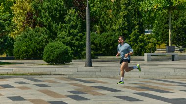 Spor giyimli ve güneş gözlüklü bir adam güneşli şehir parkında koşuyor. Fiziksel dayanıklılık, tek motivasyon, spor anlayışı, kamusal alan eğitimi ve rekabetçi ruh kavramı..