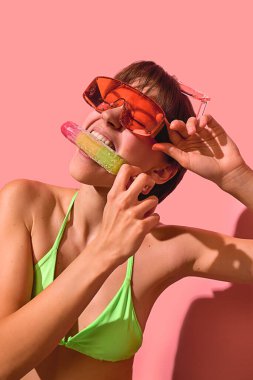 Neon bikinili neşeli kadın büyük güneş gözlüklü gökkuşağı buzlu dondurmayı ısırıyor. Yaz sıcağı kavramı, pop atıştırmalığı görseller, eğlenceli gençlik modası, canlı renkli tasarım..