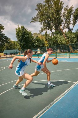 İki bayan basketbolcu bulutlu gökyüzünün altında açık bir sahada yarışıyor. Kadın sporunda yoğunluk kavramı, rekabet duygusu, gençlerin güçlendirilmesi, açık hava fitness markalaşması, basketbol kültürü