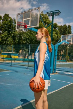 Basketbol oynayan genç bir kadın açık havada mavi gökyüzünün altında duruyor. Maçtan önce sakin konsantrasyon, spor konsantrasyonu, açık hava genç atletizmi, maç öncesi kararlılık kavramı