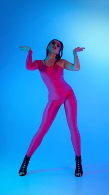 Canlı pembe beden kıyafetli dansçı mavi neon ışıkla dinamik bir şekilde poz veriyor. Agresif ritim kavramı, modern koreografi, gençlik ifadesi.