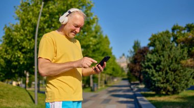 Yaşlı bir adam sabah yürüyüşünde müzik dinlerken akıllı telefonla iletişim kuruyor. Üst düzey teknoloji kullanımı, sağlıklı yaşam, sabah rutinleri, dijital sağlık ve motivasyon kavramı.