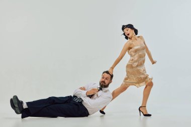 Flapper elbiseli bir kadın, elinde içkiyle yerde yatarken adamı saçından sürüklüyor. Mizahi cinsiyet rolü değişimi, retro ilişki hikaye anlatımı, alkol markalaşması, tiyatro sahneleme kavramı.