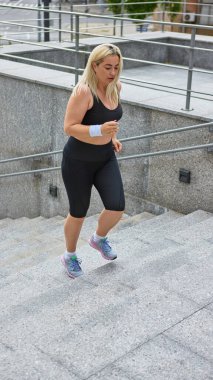 Siyah spor giyim kuşam giymiş bir kadın şehrin merdivenlerinde koşuyor. Sağlıklı yaşam tarzı, vücut pozitifliği, kentsel zindelik, aktif yaşlanma ve kadınların sağlığındaki kararlılık kavramı.