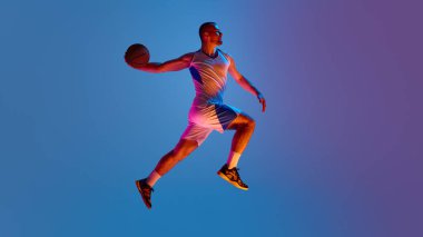 Mor ve mavi renkli arka plan ışıklandırmasıyla havada basketbol oyuncusu. Yanal momentum kavramı, erkek gücü, takım sürüşü görselleri ve gençler için stilize spor kampanyaları.