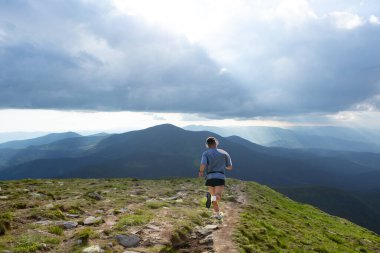 Erkek sporcu kolajı dramatik bulutlu gökyüzünün altında dağ sırtı boyunca koşuyor. Zihinsel berraklık, açık hava eğlencesi, maraton pazarlama, fitness izleme ve sağlıklı yaşam kavramı..