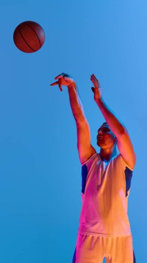 Basketbol atleti, parlak ışıklandırma ve mavi arka planla topu fırlatıyor. Hassas atış, teknik uzmanlık, spor disiplini ve editoryal kullanım için dikey eylem kavramı.