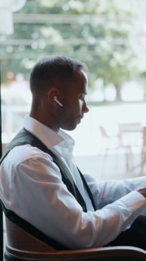 Beyaz tişörtlü bir adam büyük parlak pencerenin yanındaki kafede dizüstü bilgisayarla çalışıyor. Melez işi, iş danışmanlığı, liderlik markalaşması, uzak toplantılar ve teknoloji kavramı..