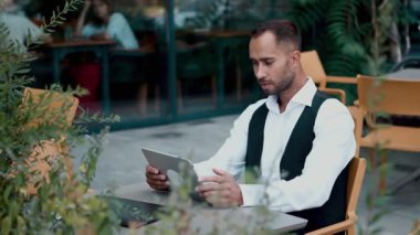 İş adamı açık masada otururken ciddi ciddi tablet okuyor. Uzaktan çalışma, iş odaklanma, çevrimiçi toplantılar, teknoloji okuryazarlığı ve açık hava iş dengesi kavramı.