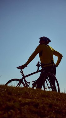 Silüetli bisikletçi gün batımında elleri kalçalarında tepenin üstünde dinleniyor. Yansıma kavramı, kişisel başarı, dayanıklılık zihniyeti, amaç ile yaşlanma, motivasyonel hikaye anlatımı.