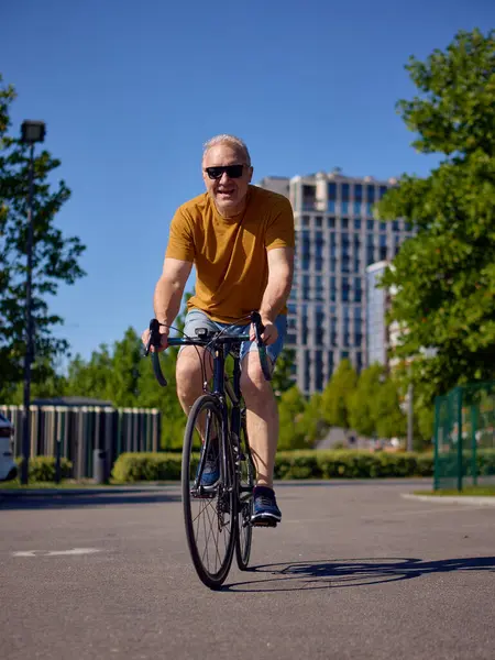 Orta yaşlı bir adam yazın şehir içinde gelişigüzel bisiklet sürüyor. Günlük fitness, kentsel refah, rahat yaşam tarzı ve sürdürülebilir şehir ulaşımı kavramı.