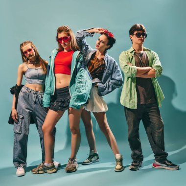 Kendine güvenen ifadelerle mavi arka planda günlük sokak kıyafetleriyle poz veren şık bir grup genç arkadaş. Gençlik kültürü, moda, yaşam tarzı ve yaratıcı reklamcılık kavramı.