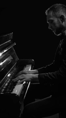 Takım elbiseli bir adamın dramatik yan ışıklandırmada piyano çalarkenki siyah beyaz portresi. Klasik müzik performansı, sahne sanatı, duygusal ifade, konser tanıtımı kavramı.