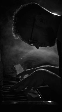 Piyanist dumanlı arka planda piyanonun üzerine derin duygularla eğiliyor. Müzik yaratıcılığı kavramı, sanatsal sahne anı, caz festivali reklamları, konser görselleri, kültürel tanıtım.