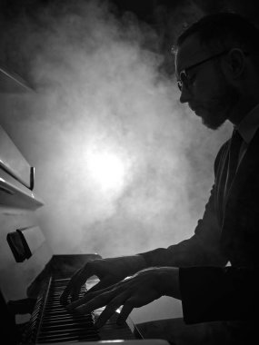 Dumanlı atmosferde piyano çalan adam odaklanmış ifadeyle. Canlı müzik performansı konsepti, konser tanıtımı, sanatsal portre, caz kulübü posteri, kültürel etkinlik reklamcılığı.