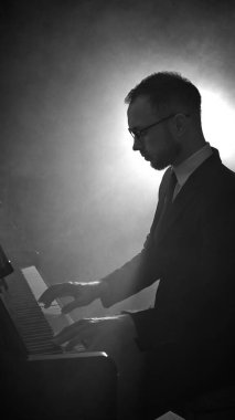 Takım elbiseli bir erkek piyanist, piyanoda, yumuşak arka plan ışığı altında derin bir konsantrasyonla performans sergiliyor. Müzikal sanatçılık kavramı, sahne performansı, kültürel tanıtım, güzel sanatlar etkinlikleri.