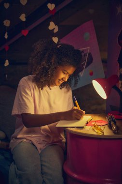 Kıvırcık saçlı genç kız kırmızı lambanın altındaki masada oturuyor, deftere retro tavan arasına odaklanmış bir şekilde yazıyor. Yaratıcılık, günlük tutma ve kişisel yansıma kavramı.
