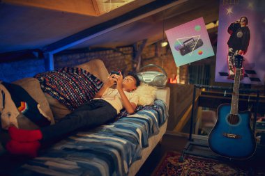 Çocuk eski dekore edilmiş tavan arasında koltukta uzanmış el bilgisayarı konsoluyla oynuyor. Oyuncu nostaljisi, gençlik kültürü ve 90 'lardan esinlenilmiş şık bir iç mekanda rahatlama kavramı.