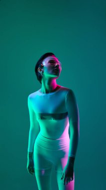 Beyaz elbiseli ve neon güneşlikli bir kadın siber ışıklandırmanın altında cyan ve morumsu tonlarda duruyor. Gelecekçi moda kavramı, modern kimlik ve kendini ifade etme..