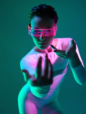 Neon rengi güneşlik ve beyaz tulumlu kadın, gök mavisi ve morumsu ışığın altında yoğun bir şekilde öne uzanıyor. Gelecekçi moda, kararlılık ve cesur bireysellik kavramı.