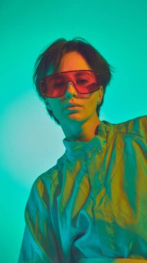 Büyük tişörtlü ve kırmızı gözlüklü bir kadın canlı deniz mavisi ışığı altında poz veriyor. Modern moda eğilimlerinde geçmişe dönük fütürizm, bireysellik ve cesur tarz ifadesi kavramı.