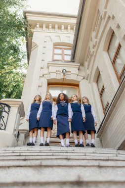 Üniformalı bir grup genç kız tarihi kampüs binasının merdivenlerinde birlikteler. Kampüs hayatı kavramı, editoryal kapak, okul topluluğu ve gençlik güçlendirme kültürü.