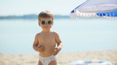 Bebek bezi takmış, güneş gözlüğü takmış kumlu sahilde duruyor. Yaz eğlencesi kavramı, çocukluk modası, açık hava yaşam tarzı, boş zaman, masumiyet, rahatlama ve aile tatili..