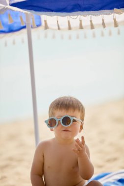 Plaj şemsiyesi altında mavi eğri güneş gözlüklü bebek. Çocukluk keyfi, yaz tatili, rahatlama, masumiyet, yaşam tarzı, aile ve rahatlık kavramı.