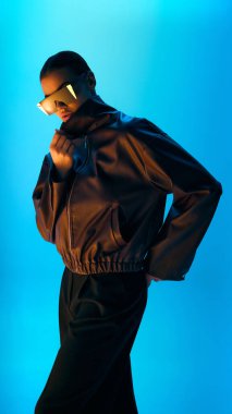 Büyük deri ceketli ve mavi ışıklı arka planda poz veren fütürist güneş gözlüklü bir model. Moda ve tarz kavramı, müzik kapakları, markalaştırıcı markalaşma ve sosyal medya görsel kodları.