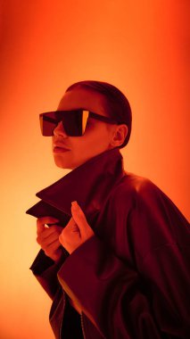 Büyük boy güneş gözlüklü ve parlak turuncu ışıklı deri ceketli kadın. Moda şıklığı, etkili kültür, dijital kimlik ve müzik kapağı tasarımı kavramı.