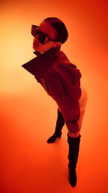 Fütürist deri ceketli, parlak turuncu arka planda büyük boy güneş gözlüklü bir kadın. Moda, bireysellik, cesur duygu, dijital sanat ve modern kültür kavramı.