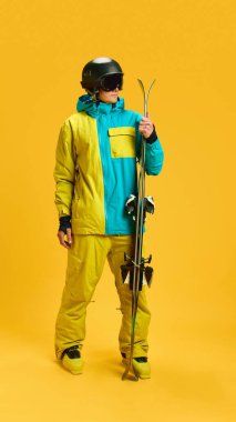 Skier holding skis upright in bright yellow studio with protective helmet and goggles. Concept of winter sports editorial, athlete portraits, training promo and seasonal gear campaigns.