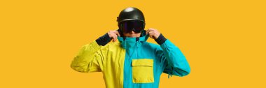 Skier adjusting helmet and goggles in yellow studio wearing ski jacket. Concept of sportswear advertising, protective gear awareness, athlete lifestyle promo and ski safety visuals.