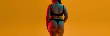 Back view of plus size woman in black lingerie against warm orange background with artistic neon glow. Concept of lingerie advertising, underwear branding, editorial campaigns, body confidence visuals