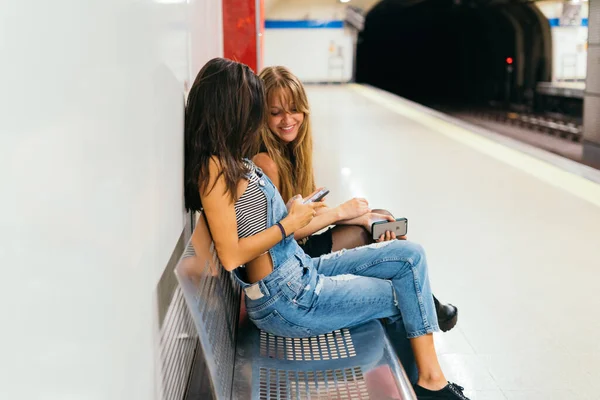 地下鉄が到着するのを待っている電話を通して見てベンチに座っている友人の女性カップル — ストック写真