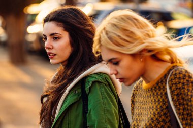 Bir çift genç bayan arkadaş kameradan başka bir yere bakarak şehirde dolaşıyorlar..