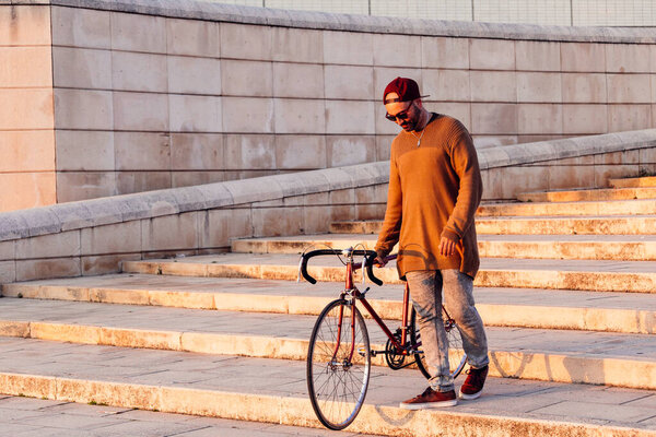 Adult man wearing a had walking down stairs in a city holding a bike.