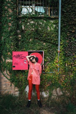 Siyah gözlüklü, kırmızı lateks pantolon ve pembe gömlekli genç bir kadın arka planda bitki örtüsü ve yangın hortumu olan bir duvarla kameraya poz veriyor..