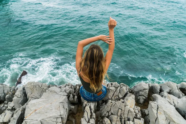 一个金发碧眼的年轻女子坐在高高的悬崖边的海面上举手的后视镜 — 图库照片