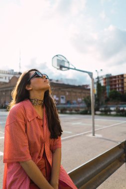 Havalı gözlüklü ve kamera önünde poz veren yetişkin bir model. Arka planda basketbol sahası olan gökyüzüne bakın.