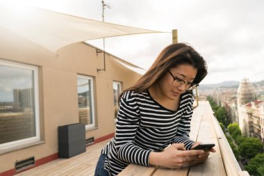 Terasta dururken akıllı telefon kullanan gözlüklü ve sıradan giyinmiş Çinli güzel bir kadın..