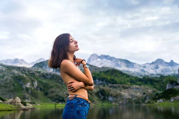 背景に湖や山の風景とカメラから離れて見て自然の中で屋外ジーンズを身に着けている若いブルネットのトップレスのサイドショット — ストック写真
